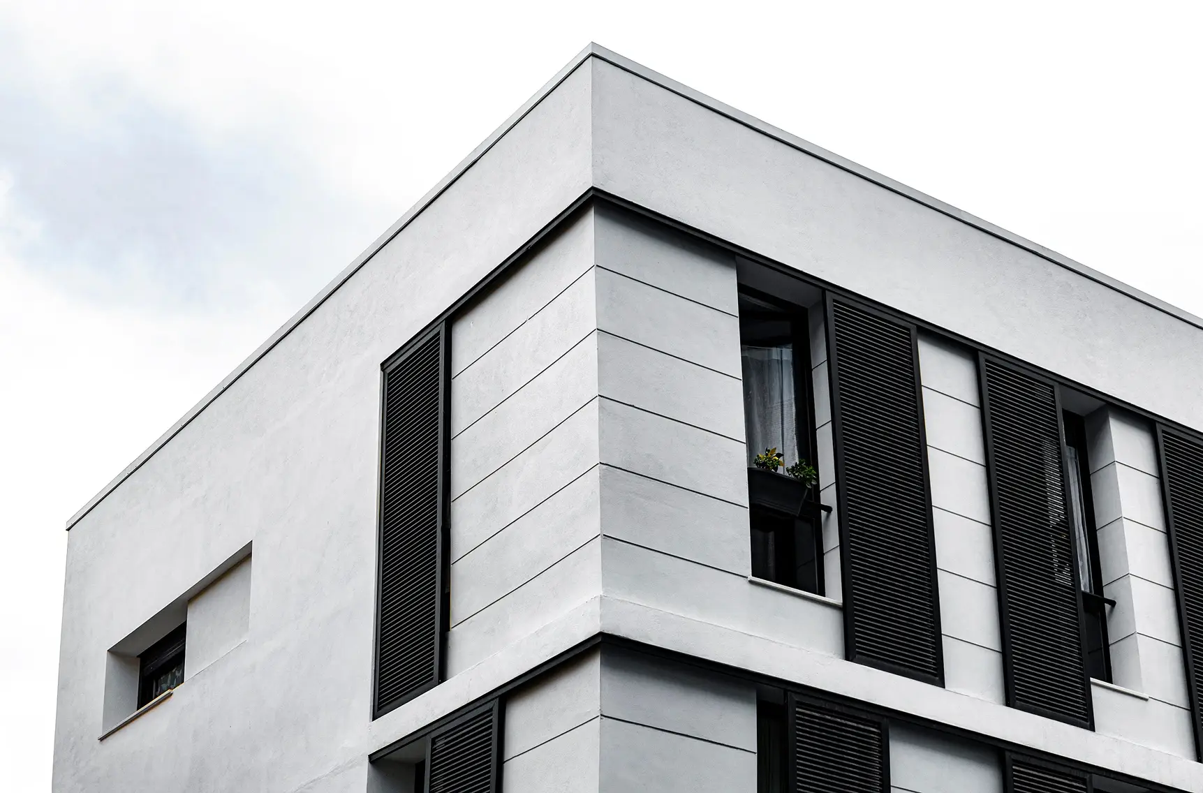 Corner of a white minimalist building featuring dark louvered shutters and clean geometric lines against a bright sky.